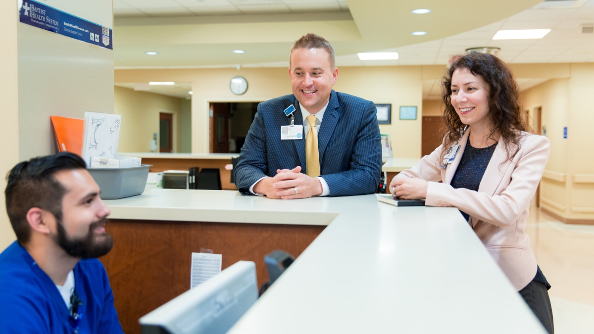 two hospital executives smile while talking to a nurse at the nurses station